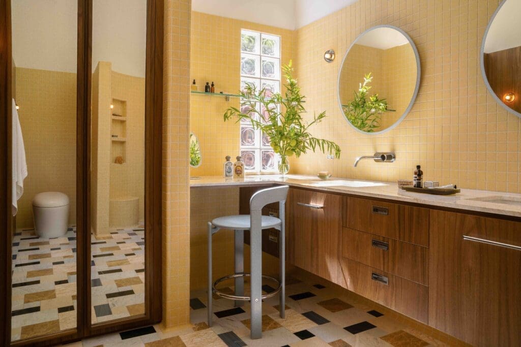 Mid-century modern bathroom designed by The Warren in Los Angeles, featuring a custom walnut vanity, round mirror, yellow tile walls, curved walk-in shower, built-in niches, and a stone mosaic marble floor.